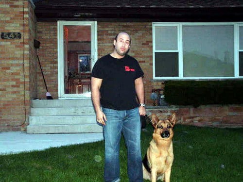 A protection Dog sitting on a grass with his owner standing beside